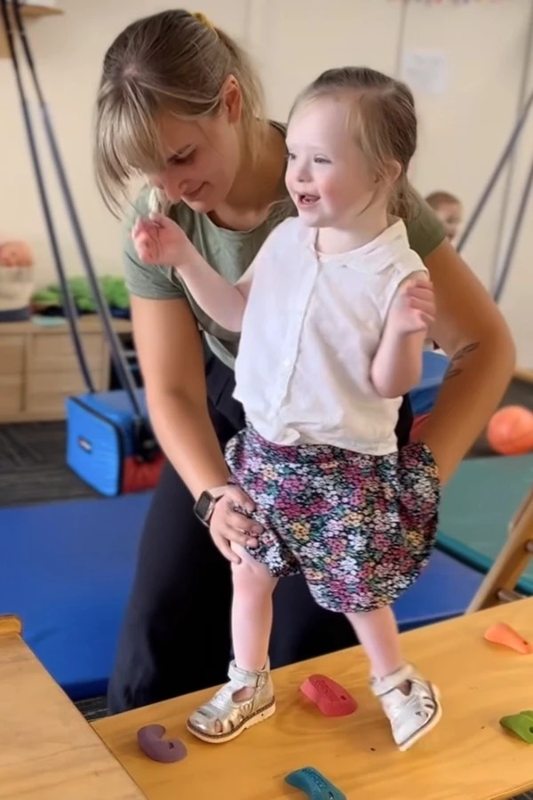 Physiotherapist helping a down syndrome girl walking on a slopped wooden board with several attached climbing knobs