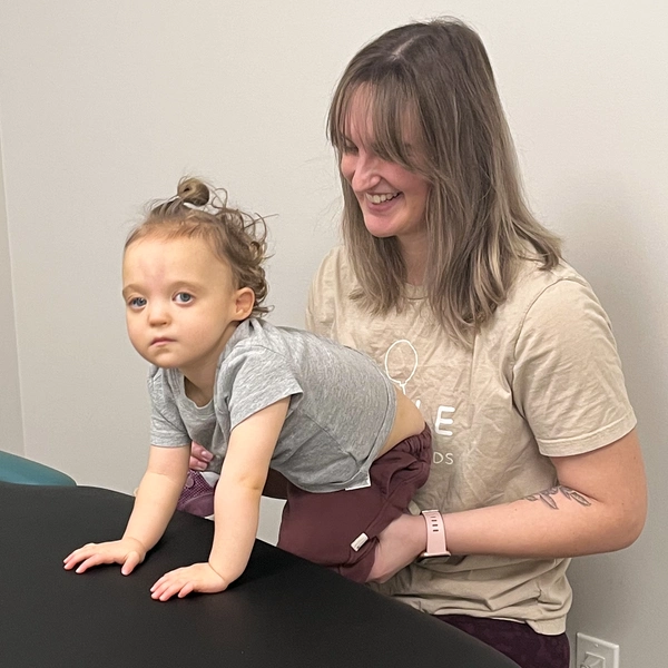 Physiotherapist holding and balancing young child with his lower legs on one hand stabilizing the upper body with the other hand, exercising Dynamic Movement Intervention while the child has his hands on a table