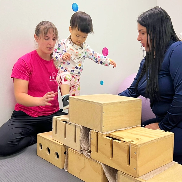 Physiotherapist holding and balancing young child walking up high wooden boxes exercising Dynamic Movement Intervention